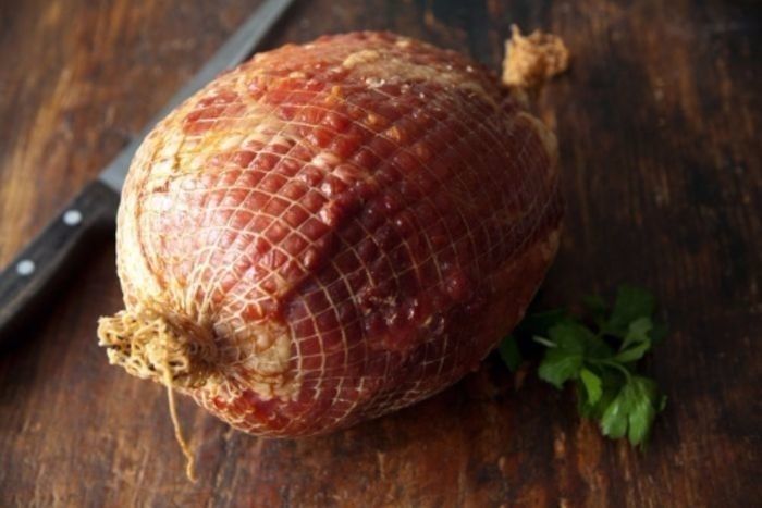 A whole smoked ham on a wooden surface, accompanied by a knife and parsley.